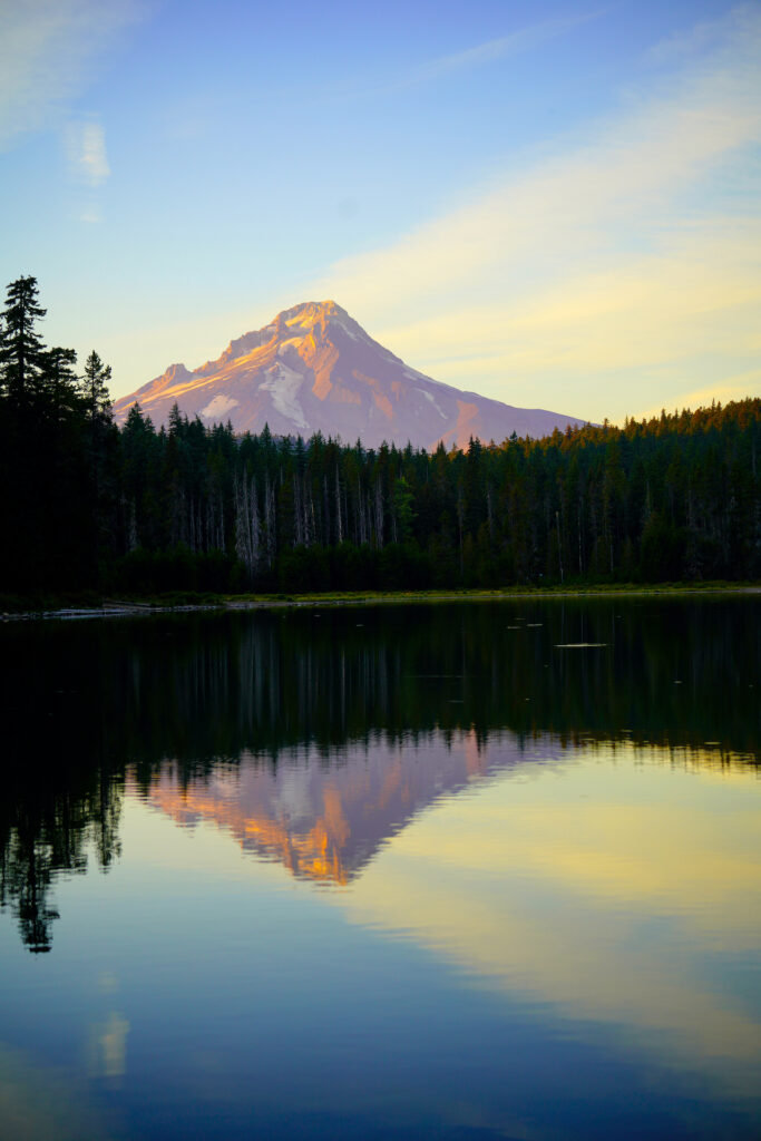 Shadows on Mt Hood take on a purple cast as wispy streaks of yellow clouds pattern the sky above it on a summer evening. The scene is reflected on the surface of Frog Lake.