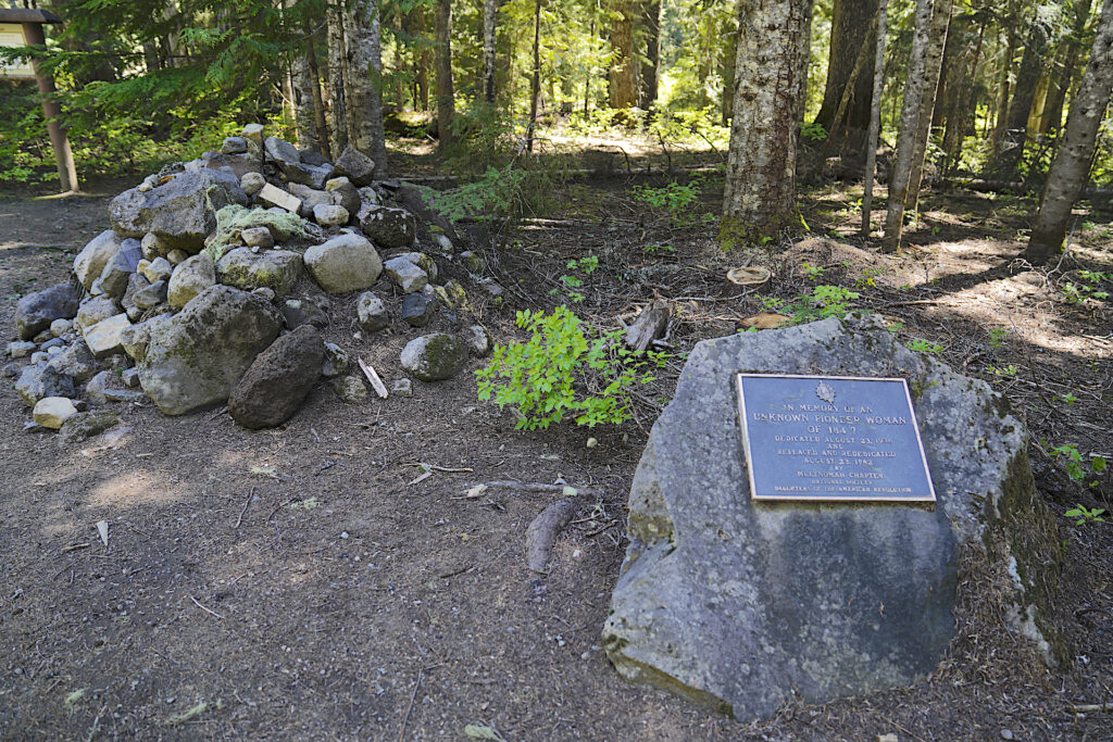 A pile of rocks marks the grave of an anonymous pioneer woman who passed away at this forested site on the slopes of Mt Hood Oregon in the 1840s. A commemorative plaque on a boulder is seen in the bottom right corner of the image.