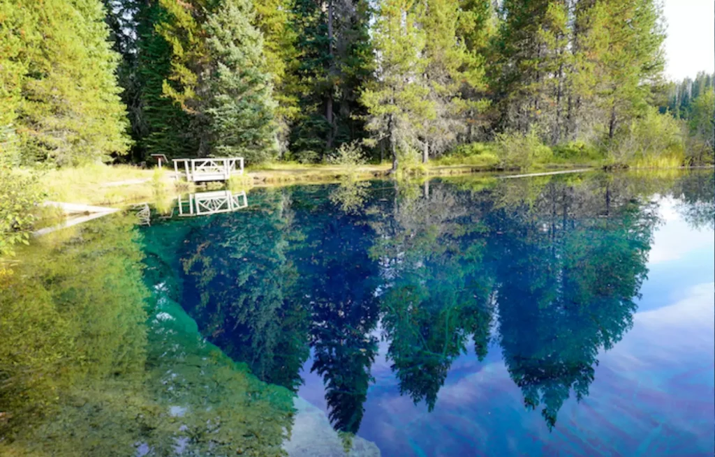 Incredibly blue and clear Little Crater Lake fills the majority of the frame, with the forested far shore at the top of the image. A little wooden viewing platform stands on the distant lake shore.