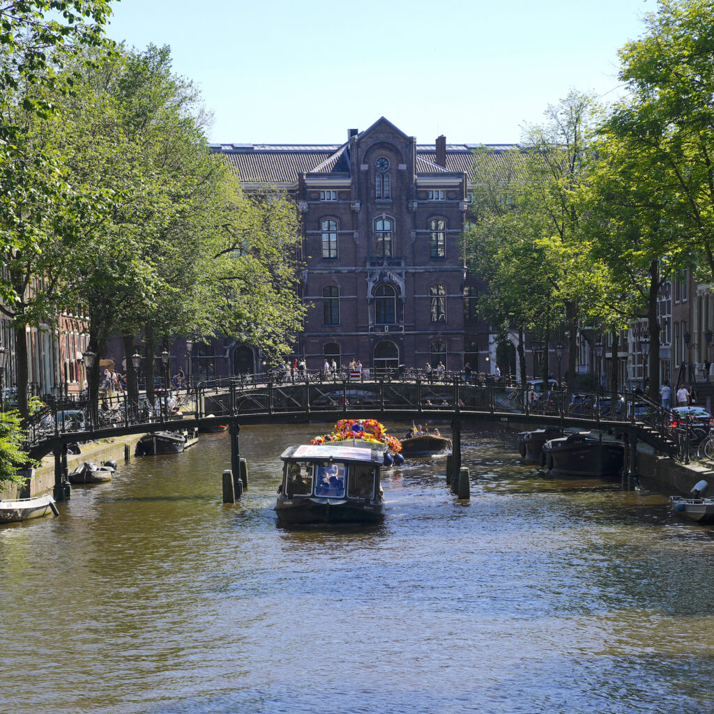 Three small boats pass under a series of footbridges as they glide up one of Amsterdam's tree-lined canals on a summer afternoon. The second boat in the line is covered in brightly colored flowers. A large historic brick building is in the background.