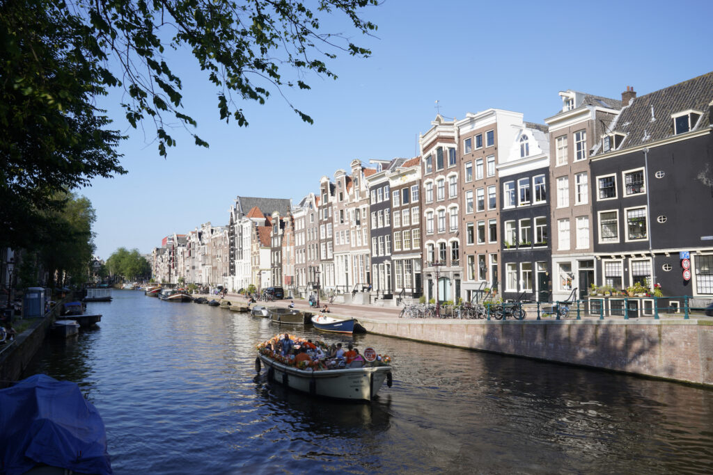 A tour boat filled with visitors plies the waters of one of the West Canals in vibrant Amsterdam. A row of tall houses lines the canal.