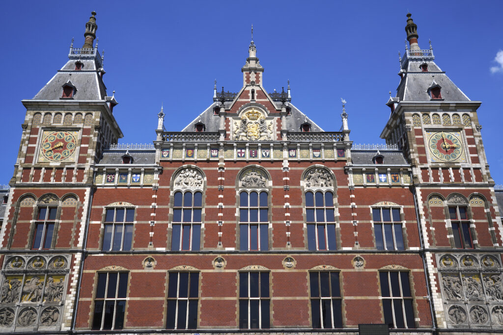 The center facade of Amsterdam Centraal Station shows off it's ornate details and red brick walls topped by a pair of clock towers on a clear summer day.