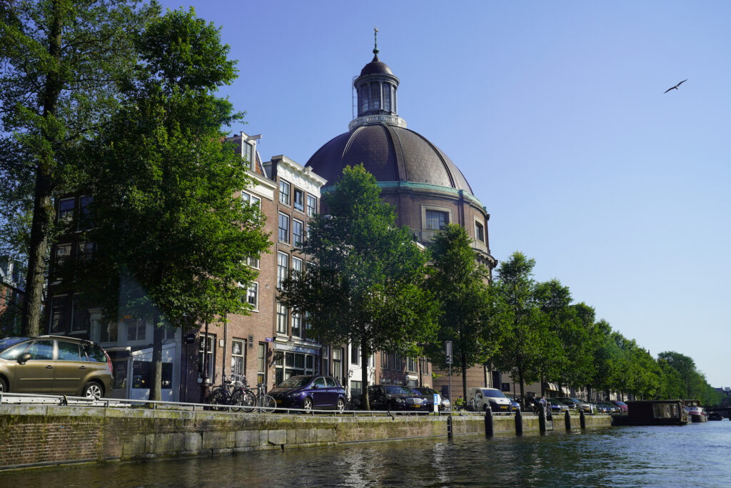 A historic building with a big domed roof stands along a tree-lined canal in Amsterdam.