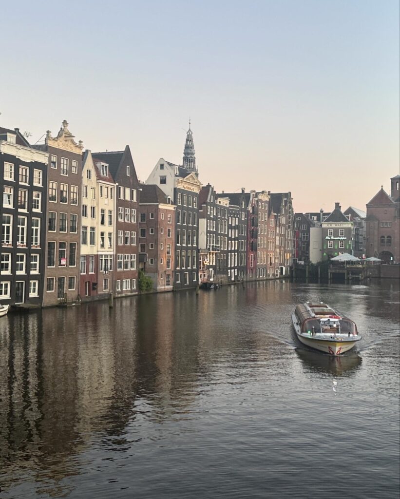 Evening glow fades on a line of waterside canal houses in Amsterdam. A glass-roofed tour boat passes by on the right.