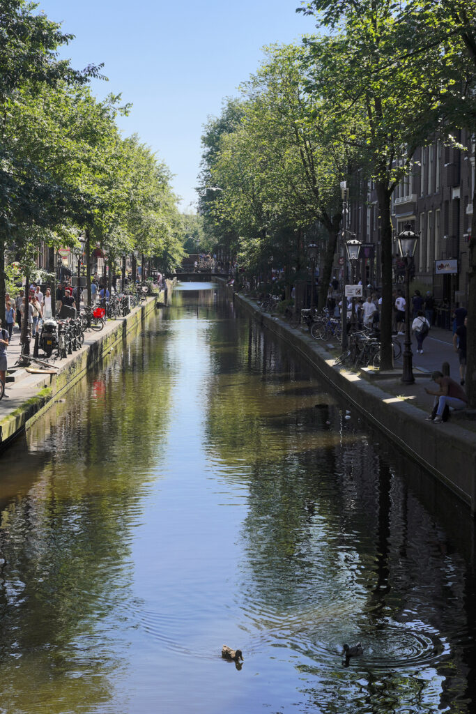 Looking straight down a slender canal in Amsterdam on a sunny summer afternoon. The tree-lined streets on either side of the canal are busy with pedestrians.