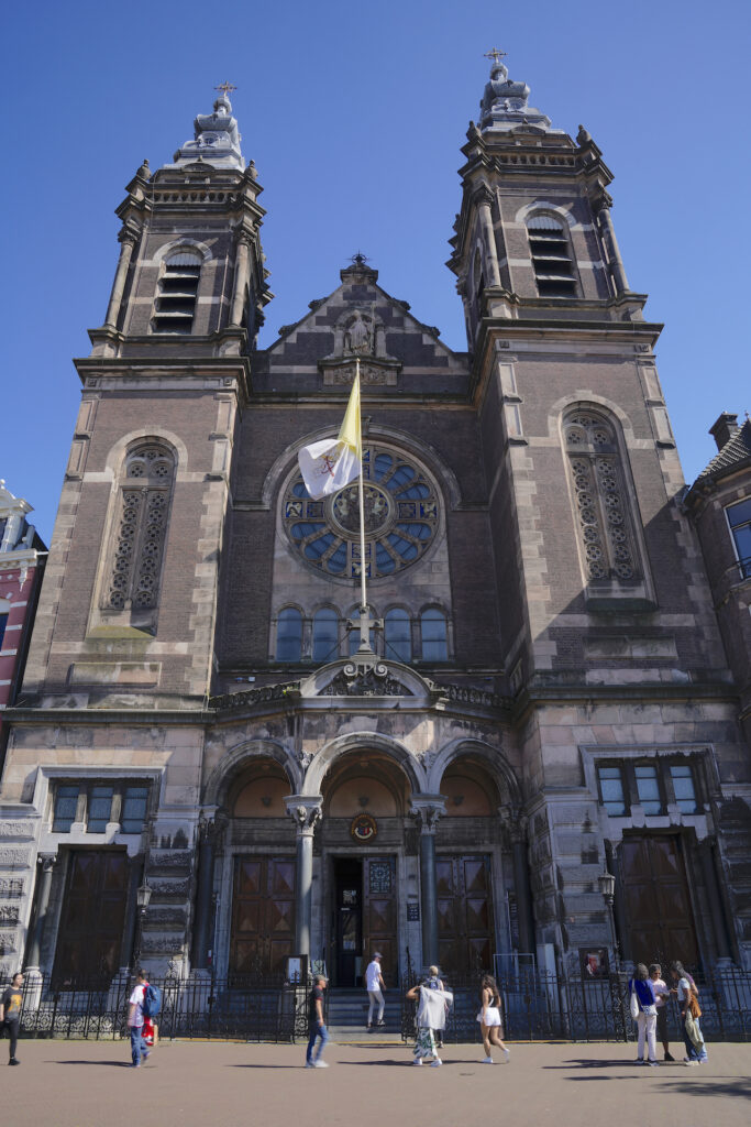 The exterior of Basilica of St Nicholas in central Amsterdam features two tall towers framing the cathedral's main doors and rose window.