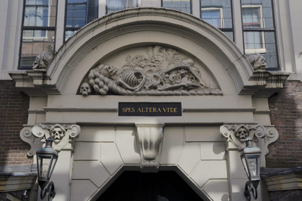 A relief sculpture of a reclining skeleton is posed above a door to the Basilica of St Nicholas in Amsterdam. A plaque is beneath it which reads "Spes Altera Vitae" meaning "hope of another life" - a reference to reaching heaven in the afterlife.