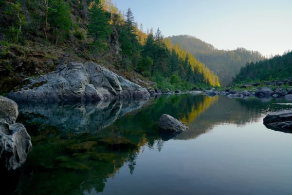 The surface of a deep clear pool in the Illinois River reflects the rocks and forest on the shore on a gentle summer evening.