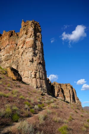 A trail leads up a grassy desert slope to the base of a tall pinnacle of basalt that protrudes from the nearby cliffs.