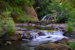 View looking up a small river with a small ledge waterfall in the foreground and a larger waterfall in the background.