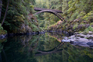 An arching foot bridge is reflected in the calm water of East Fork Lewis River in Moulton Falls Regional Park.