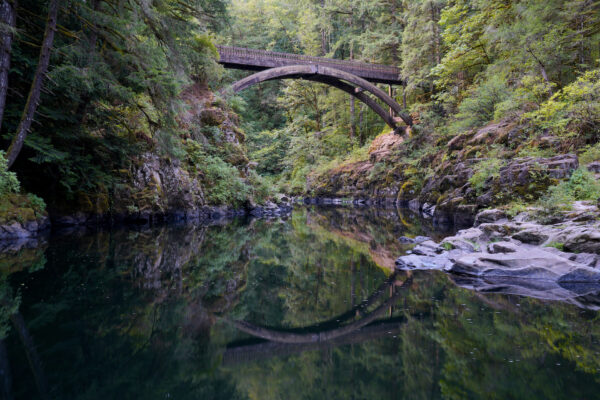 An arching foot bridge is reflected in the calm water of East Fork Lewis River in Moulton Falls Regional Park.