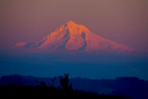 Snow-covered Mt Hood, Oregon glows in orange sunset light with layers of shadowy ridges below.