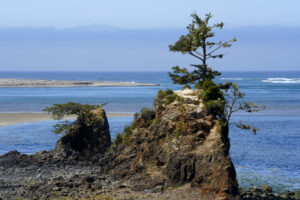 A group of two rocks, both topped with pine trees, stand in front of Siletz Bay on the Oregon Coast.