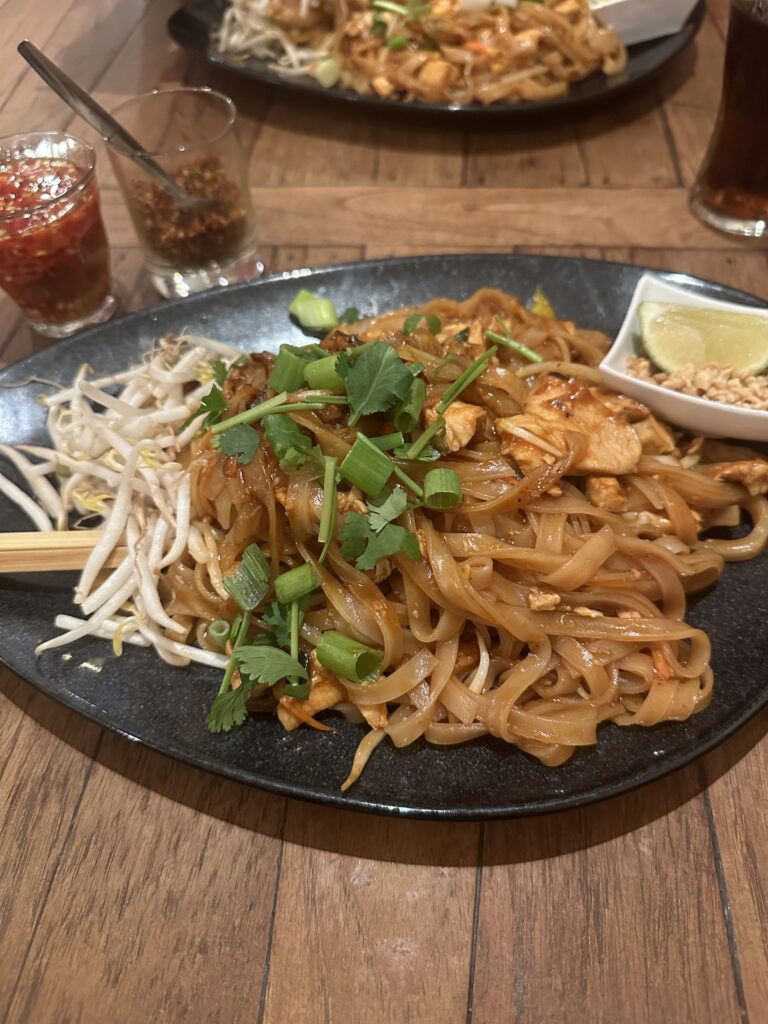 A plate of Pad Thai topped with green onions and cilantro, with fresh bean sprouts on the side and a little dish of ground peanuts and a lime wedge, accompanied by two little glasses of spicy condiments.