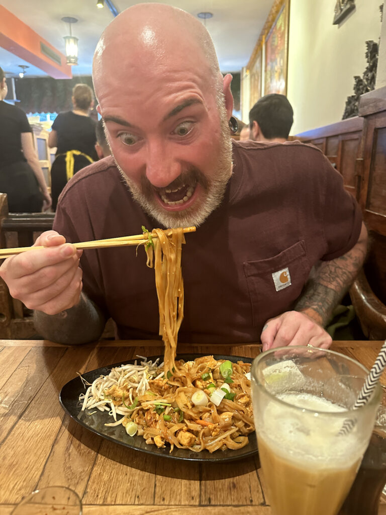 A funny picture of a wide-eyed man about to ravenously bite into the Pad Thai noodles dangling from his chopsticks.