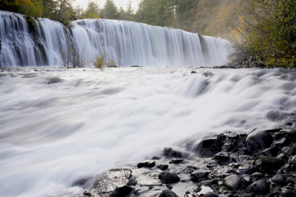 A very wide waterfall drops as a nearly solid sheet over a low cliff, then rushes downstream.