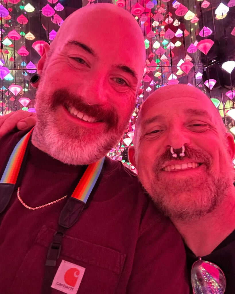 Two men pose for a selfie inside Diamond Matrix at moco Museum. The light is dark pink.