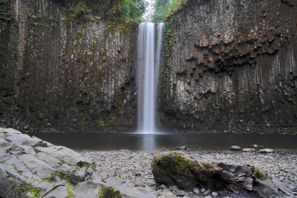 A head-on view of Abiqua Falls dropping over a gorgeous columnar basalt cliff into it's big pool, with a pebble beach and a few mossy boulders in the foreground.