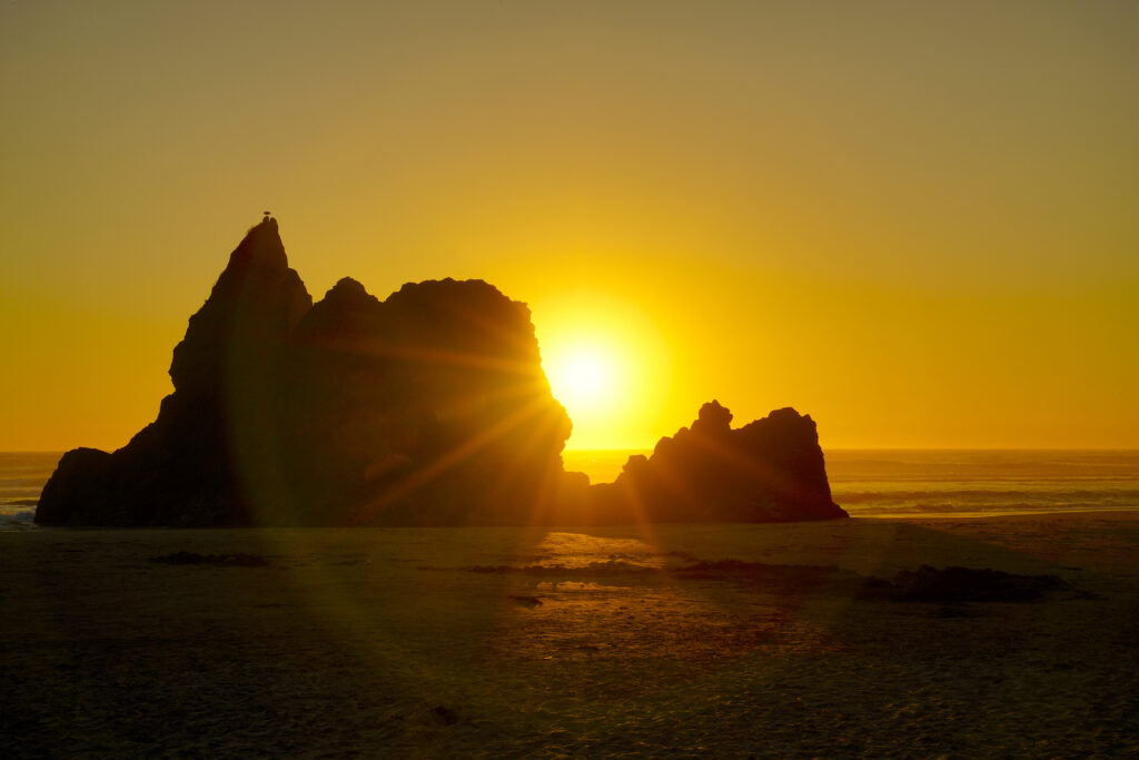 The sunset light shines through a notch in a large, craggy monolith on the beach at Arcadia State Park in Oregon.