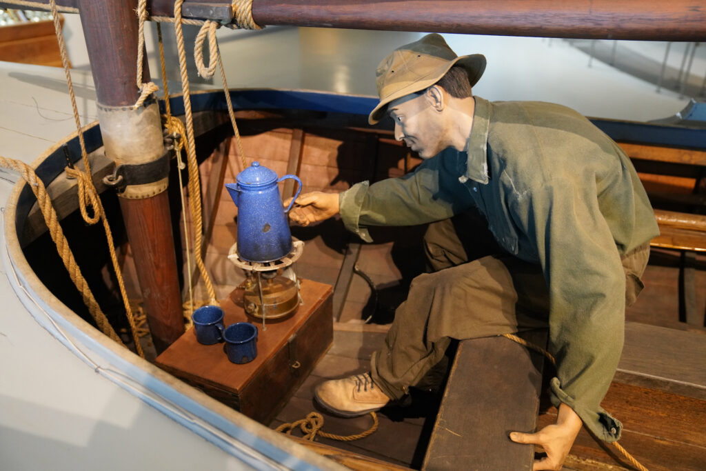 A mannequin is posed as a fisherman on a small sailboat. He's in the wooden cockpit making coffee on a portable burner.