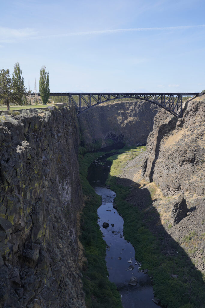 This photo looks down a desert canyon with a river flowing in the bottom. An arch bridge spans the canyon in the distance. The walls are vertical and the terrain above the canyon is flat with a few trees on the far left of the image.