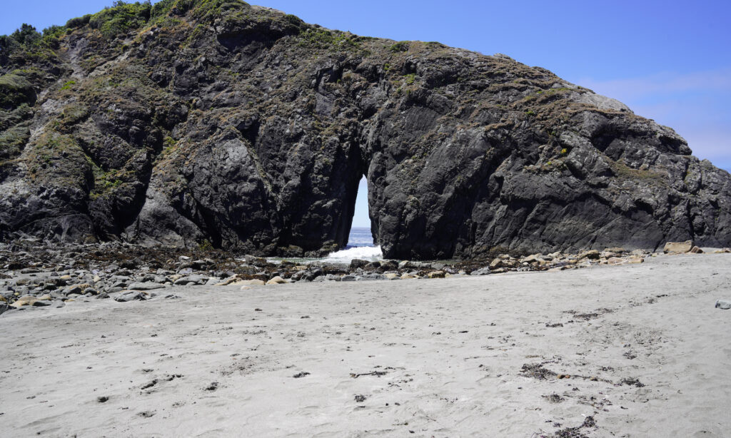 Waves crash through a wedge-shaped arch in a huge rounded megalith at Harris Beach State Park.