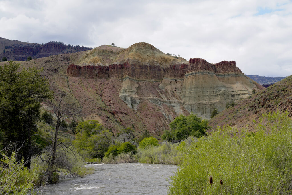 The John Day River flows toward the base of Cathedral Rock which is decorated in layered stripes of claystones in shades of rust, tan, and seafoam green.