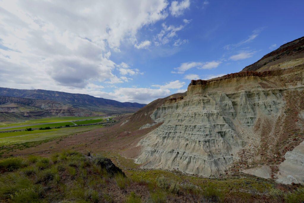 A green claystone cliff face erodes in badlands-style formations on the Flood of Fire Trail at Foree. A desert grassland environment stretches into the distance below it.