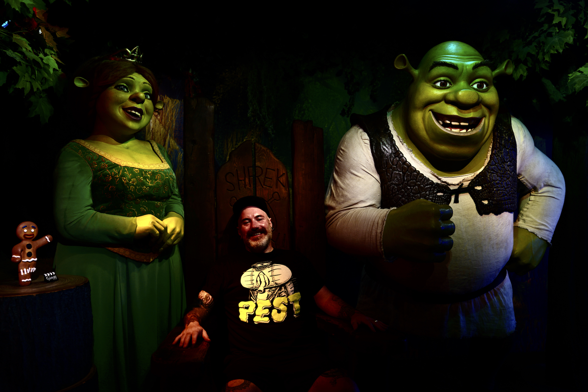 A man in a black T-shirt is seated between two large wax figures of Shrek and Fiona.