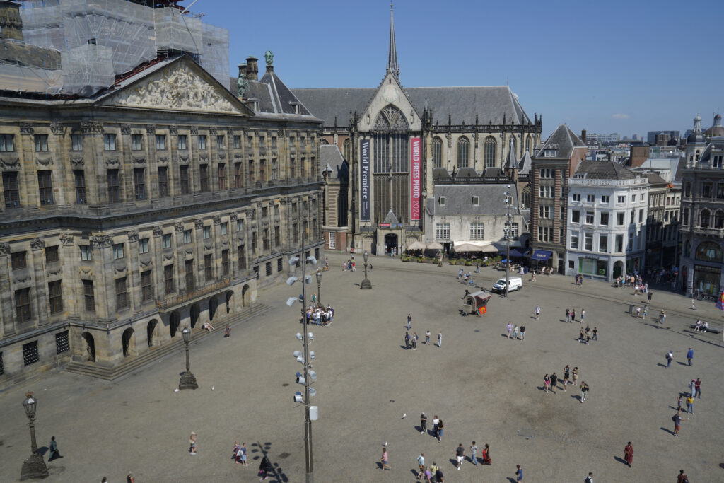 View looking down on Dam Square, where a few groups of people are sightseeing. The Royal Palace is on the left of the square and de Nieuwe Kerk (the New Church) stands at the back of the square.