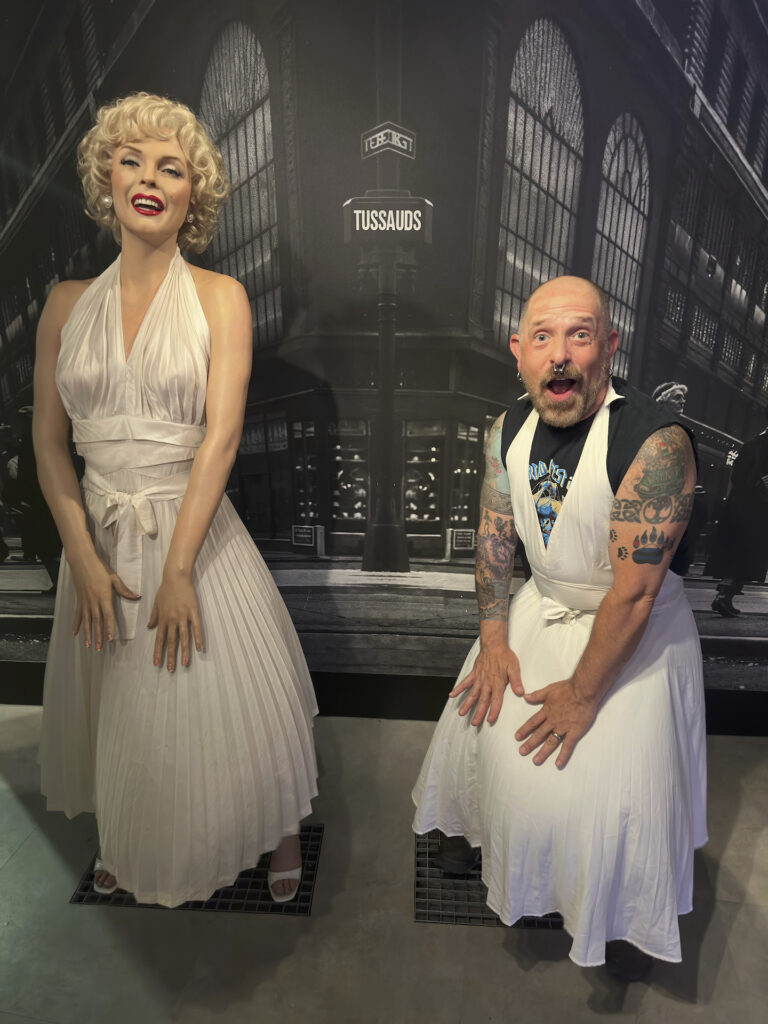 A realistic wax figure of Marilyn Monroe in her classic blowing white dress pose stands on the left, with a shorter man with a matching dress over his clothes imitating her pose on the right. A black and white photo of a 1950's era cityscape fills the background.