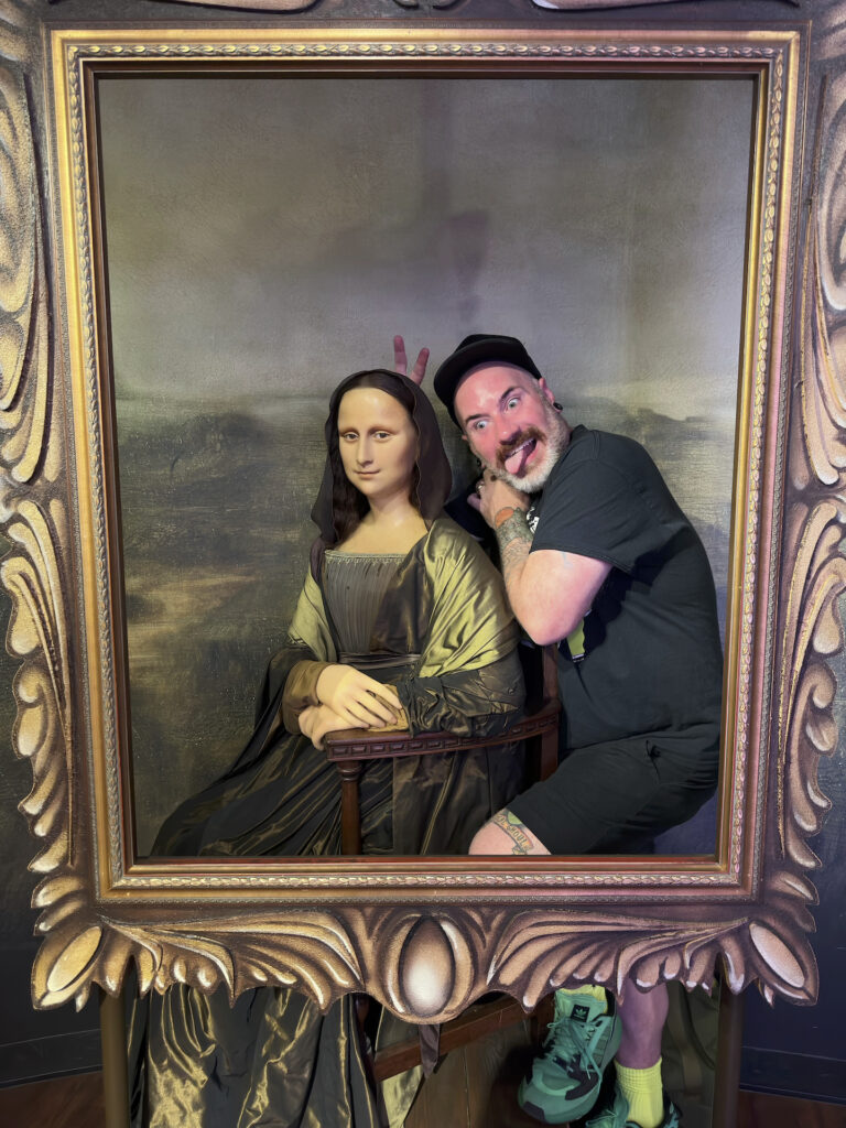 A man appears to be in the painting with Mona Lisa. He is making a silly face with tongue out and has "bunny ears" fingers raised behind her head.