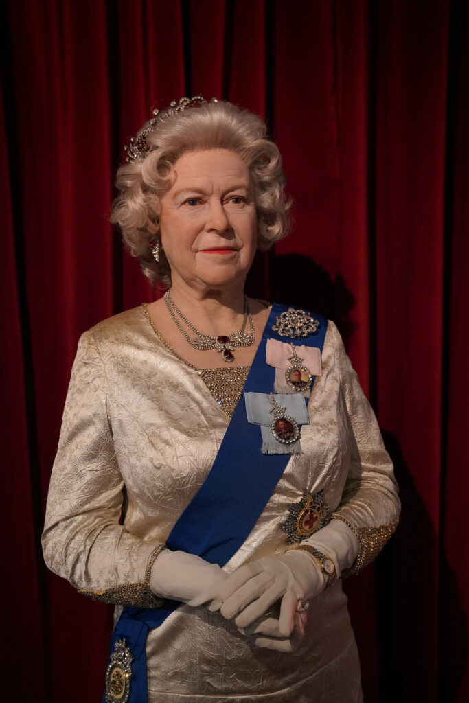 A realistic wax figure of Queen Elizabeth II of England stands in front of a red velvet curtain.