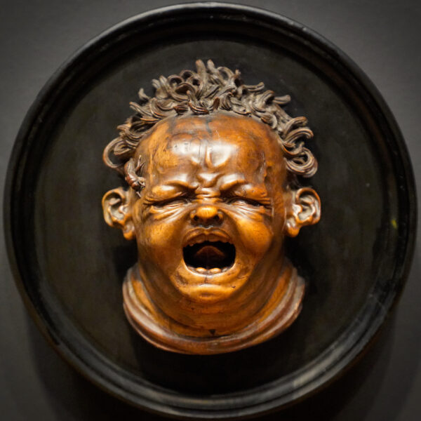 A historic wood carving of the face of a crying infant - poor guy is being stung by a bee on it's left forehead!