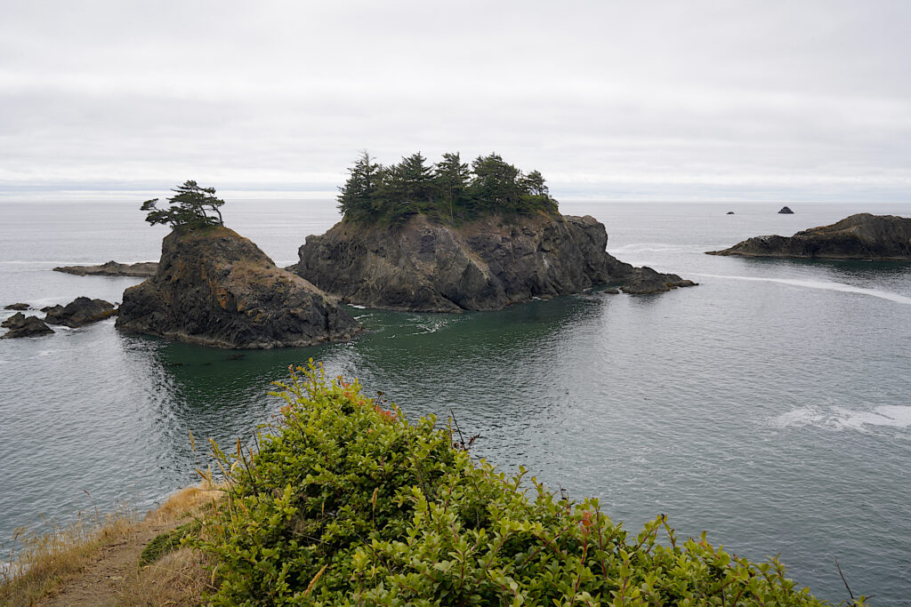 A group of large offshore rocks capped with conifers rises from calm seas on a cloudy day at Thunder Rock Cove in Samuel H. Boardman State Scenic Corridor.