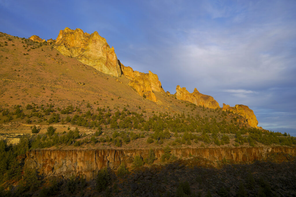 Golden sunset light shines brightly onto Staender Ridge, which rises from right to left diagonally across the image with several fins of yellow rock protruding from the ridgeline. Above the ridge the sky is blue with a cluster of nondescript clouds on the right. A line of cliffs extends across the bottom of the image with deep shadow beneath them and scattered small pine trees on top.