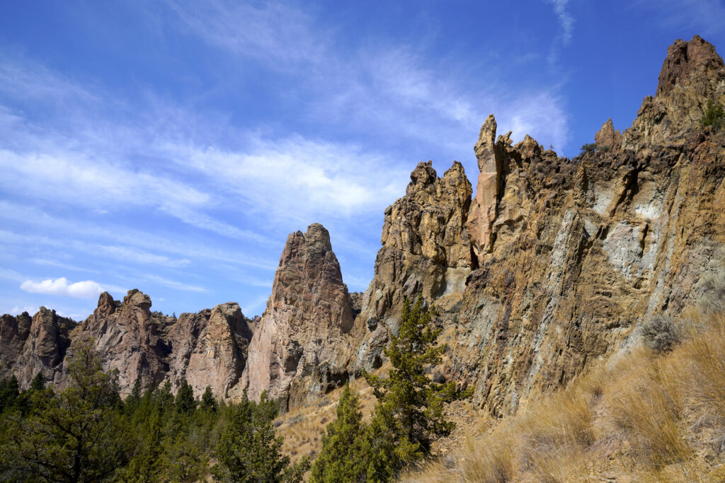 A line of pinnacles and jagged rock formations is features in this image, swooping across and up to the right corner. A blue sky with thin stripes and patches of clouds fills the top of the image, and a group of pine trees fills the lower left corner beneath the pinnacles.