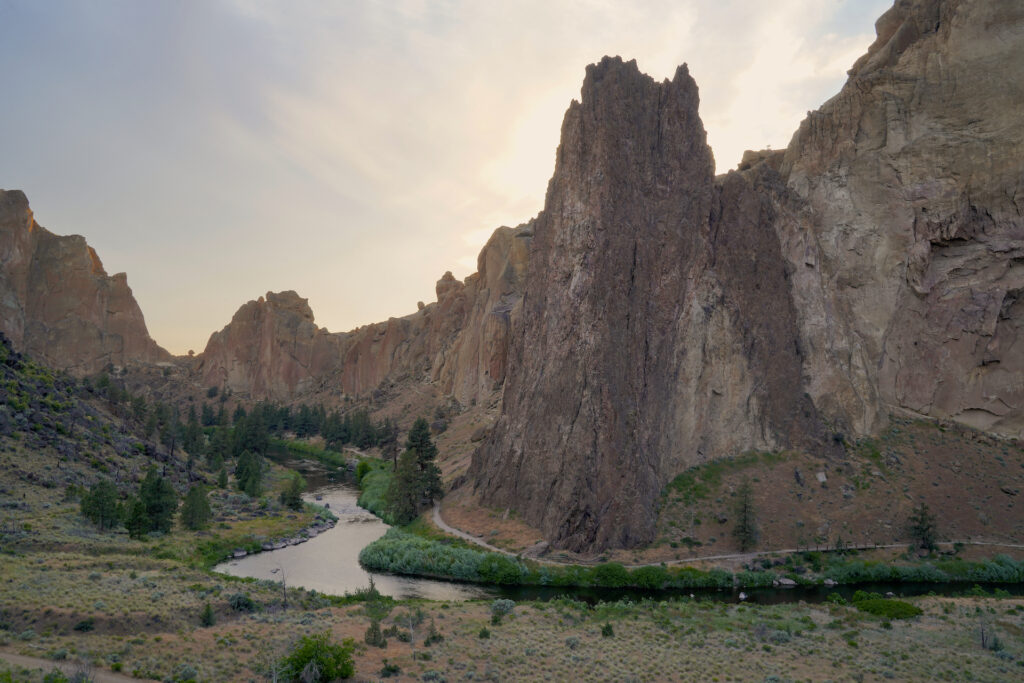 Thin clouds glow with warm evening light behind the craggy ridgeline and hulking desert cliffs of Smith Rock. A large pinnacle is the focal point, standing slightly right of center in the image. The Crooked River winds along the base of the cliffs, flanked with green vegetation and trees in the otherwise desert landscape. A trail is visible following the far shore of the river against the cliffs.