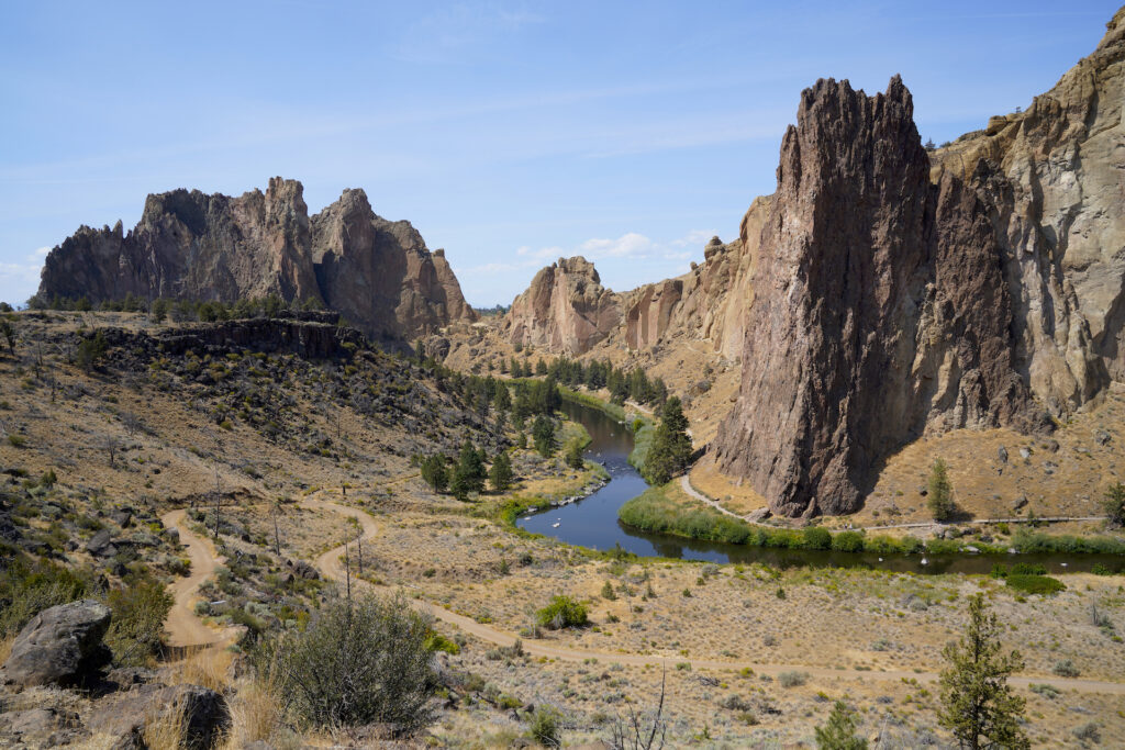 This photo features a craggy line of cliffs with serated tops which swings back into the distance in the left half of the image. A small river traces this shape at the foot of the cliffs, with greenery on it's shores. Overall the landscape is parched sagebrush desert. A wide trail, nearly a road, parallels the river on the near shore before swinging back uphill in the bottom left of the image. The sky above the cliffs is light blue with a few streaks of cloud.