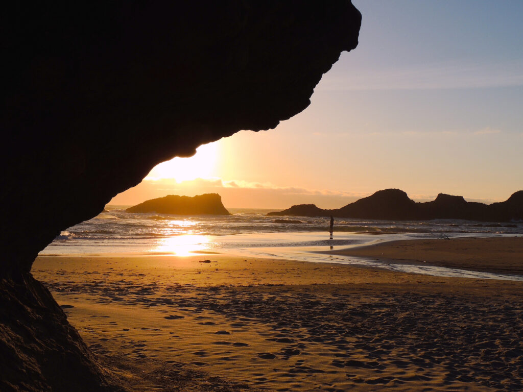 A man stands on the edge of the waves contemplating the sunset at Seal Rock Beach. A large rock overhangs the view in the upper left corner.