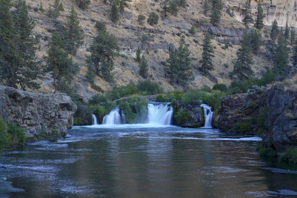 View looking upstream at a river dropping 20 feet over a wide ledge in four channels (center right is largest, far left is smallest). Desert canyon walls, sparsely covered with pine trees, rise behind the falls, filling the image. A large pool fills the bottom of the image with low, rocky cliffs on the sides. It is a peaceful summer evening and the light is soft.