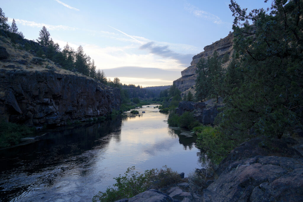 This photo looks down the Deschutes River in Central Oregon as is gently flows through a desert canyon on a peaceful summer evening. Grey and cream-colored bands of clouds near the horizon and the blue sky above are reflected by the water. A large juniper tree growing from a group of rocks fills the right edge of the image.