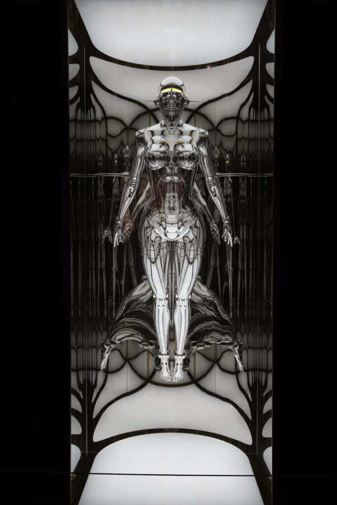 A futuristic reflective metal artwork of a robotic-humanoid figure with yellow eyes appears to flat inside a glass box with glowing white lighting. It is called "untitled_Sexy Robot Type II floating" and was created by Hajime Sorayama.