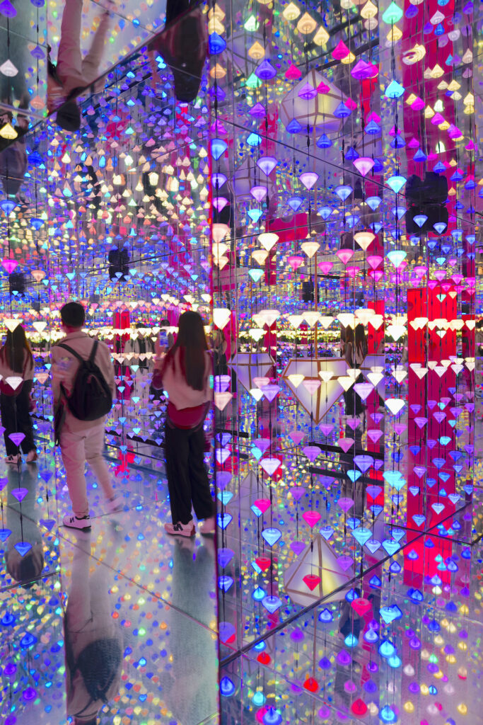 A pair of visitors get lost inside Diamond Matrix at moco Museum Amsterdam, where mirrored walls and ceilings reflect the constantly changing colors of hundreds of vertically-strung diamond-shaped lights.