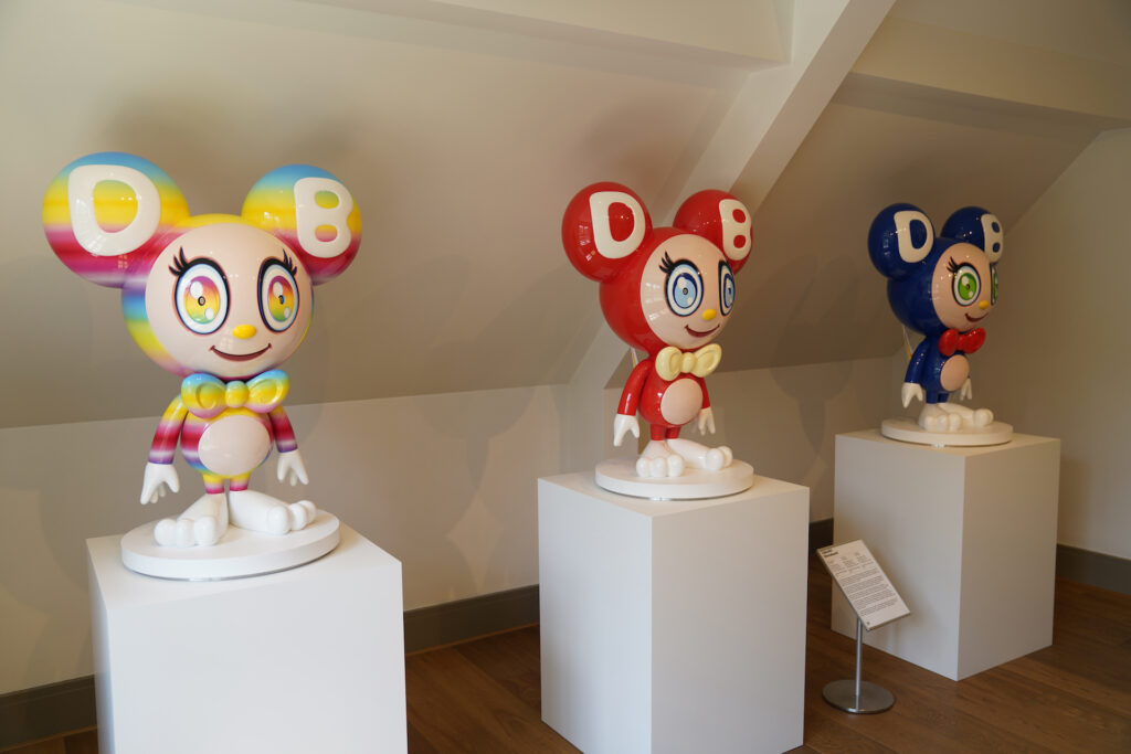 Three plastic statues, all variations of the same idea (a cartoonish figure with round bubble ears, big eyes, a colorful bodies) stand on white pedastals inside moco Museum.