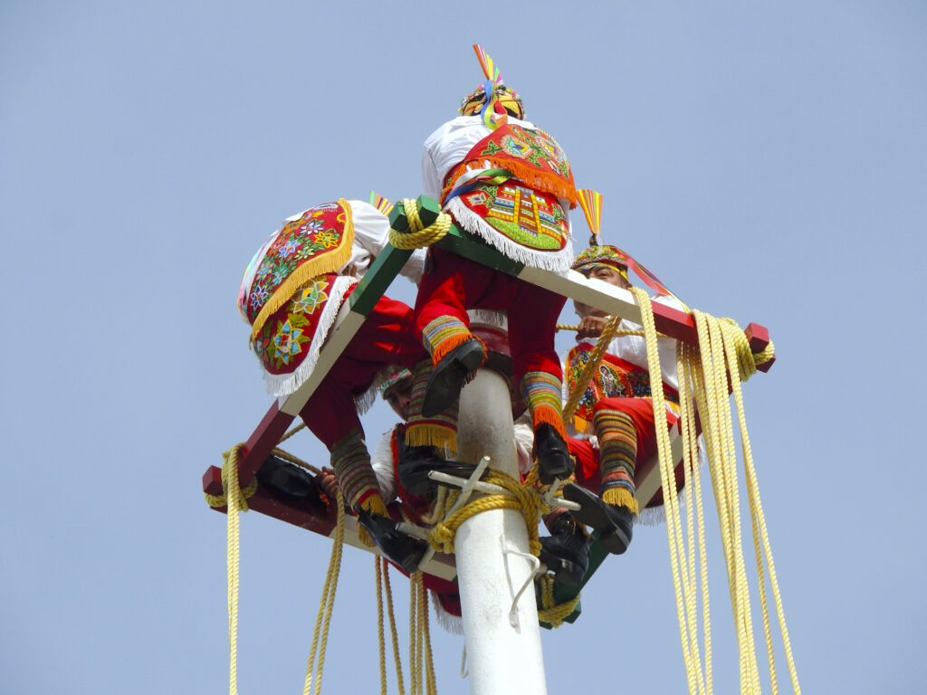 Four men in colorful traditional costumes sit on a small platform at the top of a tall pole with ropes hanging down below them. They are about to perform a traditional Mesoamerican ceremony known as "Danza de los Valodores".