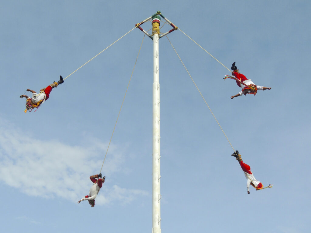 Four men in bright traditional costumes swing upside down on ropes connected to the top of a tall pole in a Mesoamerican ceremony known as "Danza de los Valodores".