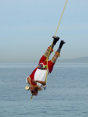 A performer of the traditional Mesoamerican ceremony known as "Danza de los Valodores" is suspended upside down from a rope as he swings through the air. This man is the musician; in one hand he plays a small drum with a stick while the other plays the notes on the small whistle he is blowing through.