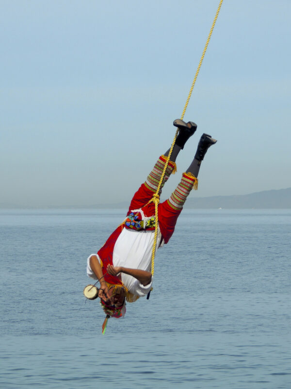 A performer of the traditional Mesoamerican ceremony known as "Danza de los Valodores" is suspended upside down from a rope as he swings through the air. This man is the musician; in one hand he plays a small drum with a stick while the other plays the notes on the small whistle he is blowing through.